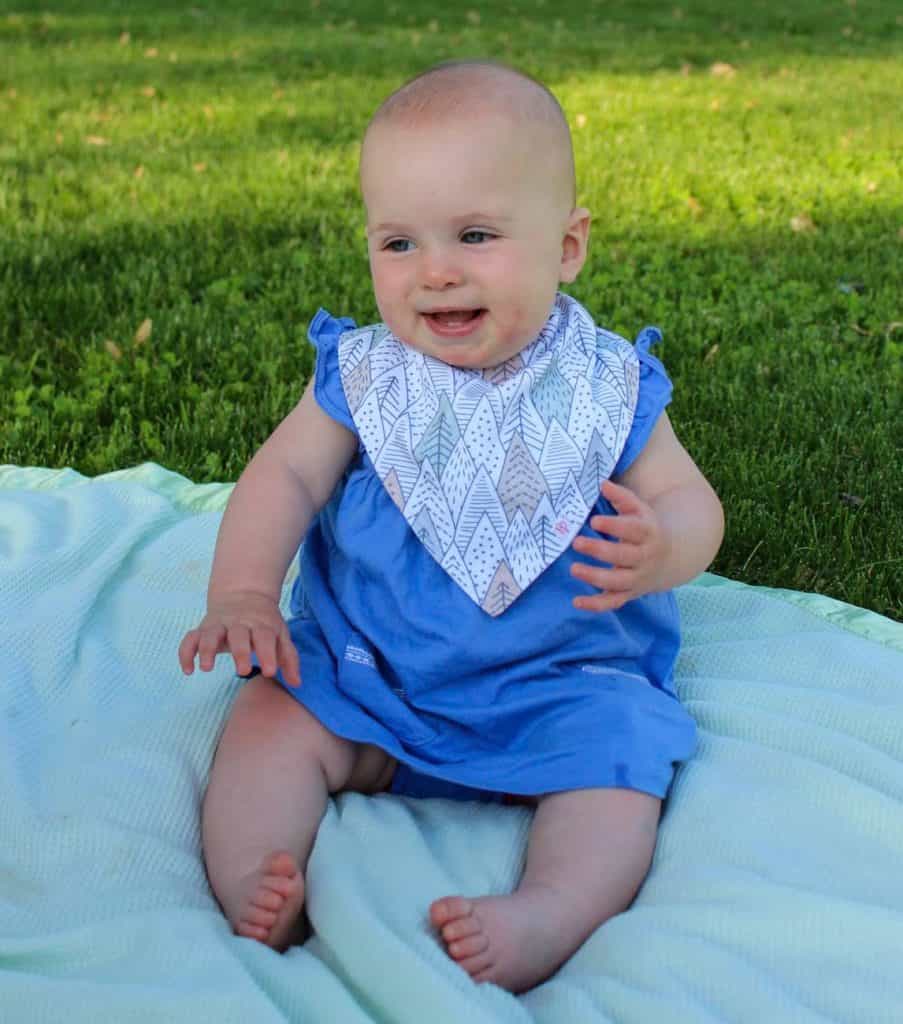 baby in blue dress and drool bib sitting on blankets on grass