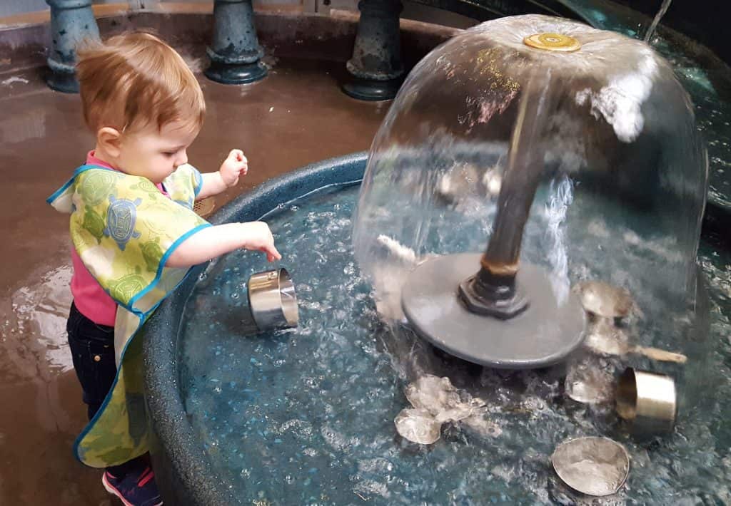 toddler playing in a water feature with measuring cups