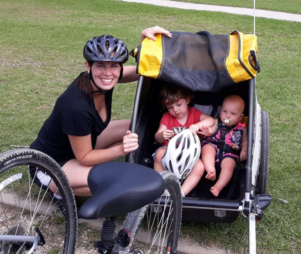 Woman in helmet next to a baby and toddler sitting in a Burley bike trailer
