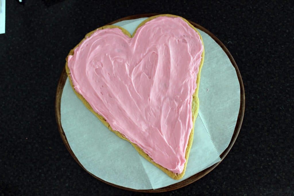 large heart shaped sugar cookie on pizza stone covered in pink cream cheese frosting