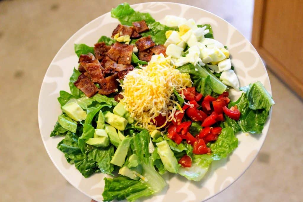 a plate of Cobb salad with toppings in sections; bacon, avocado, hard boiled egg, tomato, cheese, and bacon