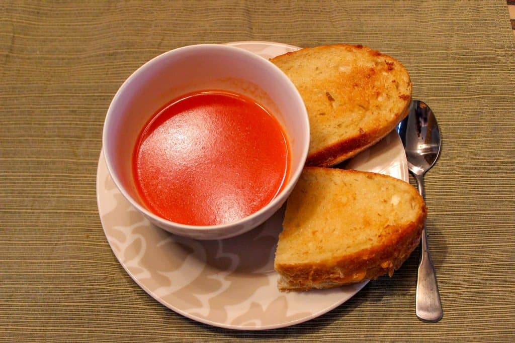 A bowl of tomato soup on a plate with a grilled cheese sandwich