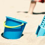 sand pail and shovel stuck in sand and a child's legs walking on the beach