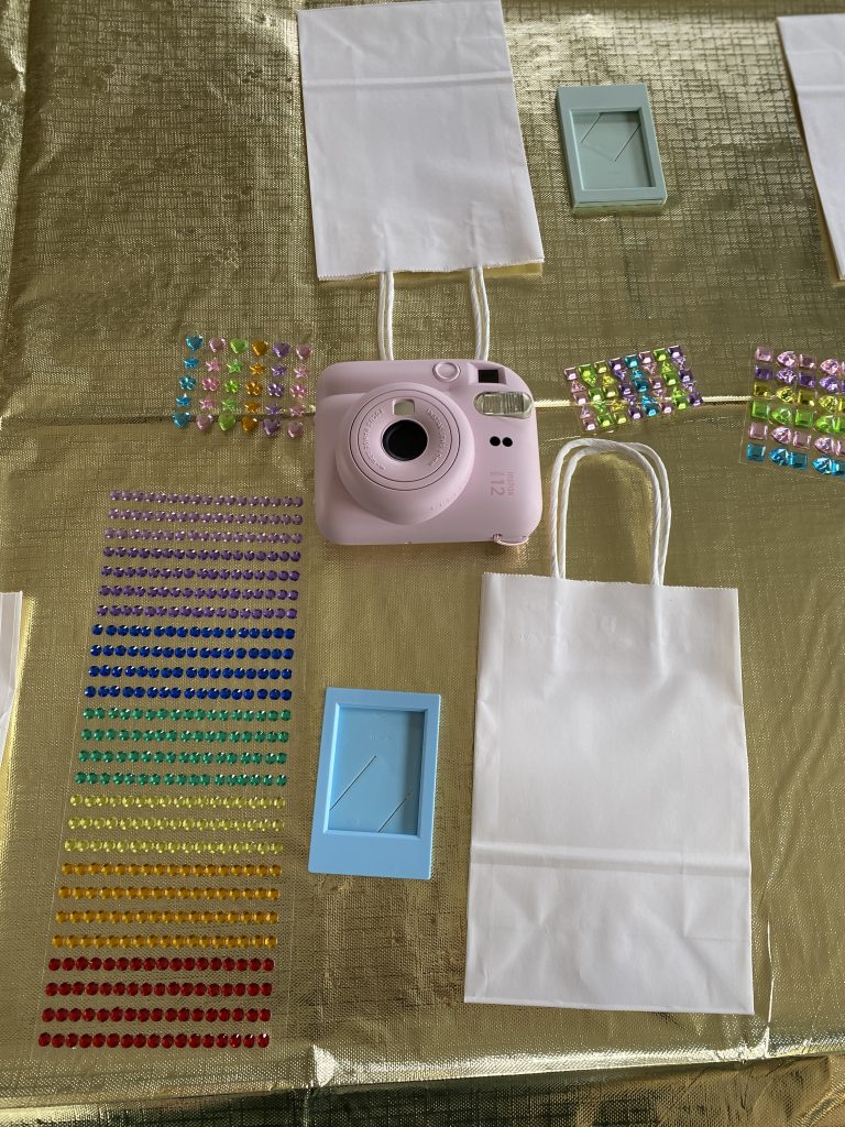 Blank white favor bags ready to be decorated laying on the table with gem stickers, small picture frame, and Instax Mini camera
